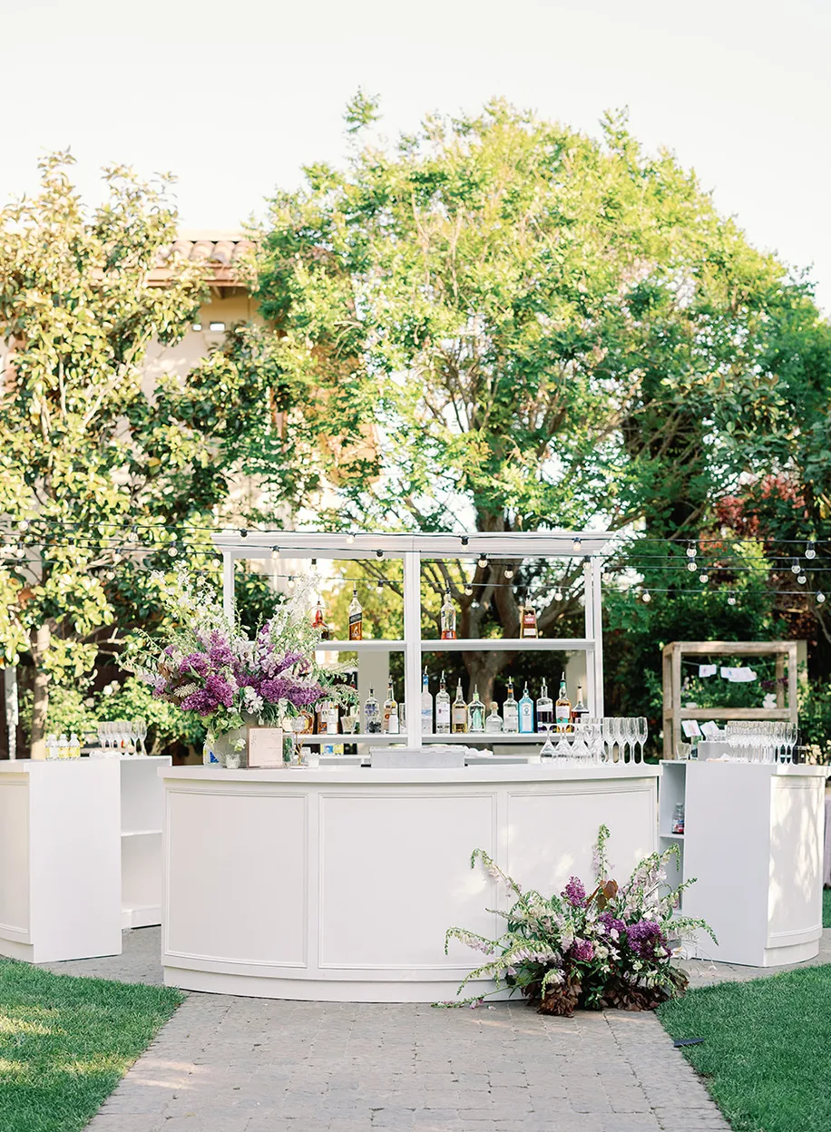Outdoor cocktail bar with a curved white counter set among lush green trees on a sunny day