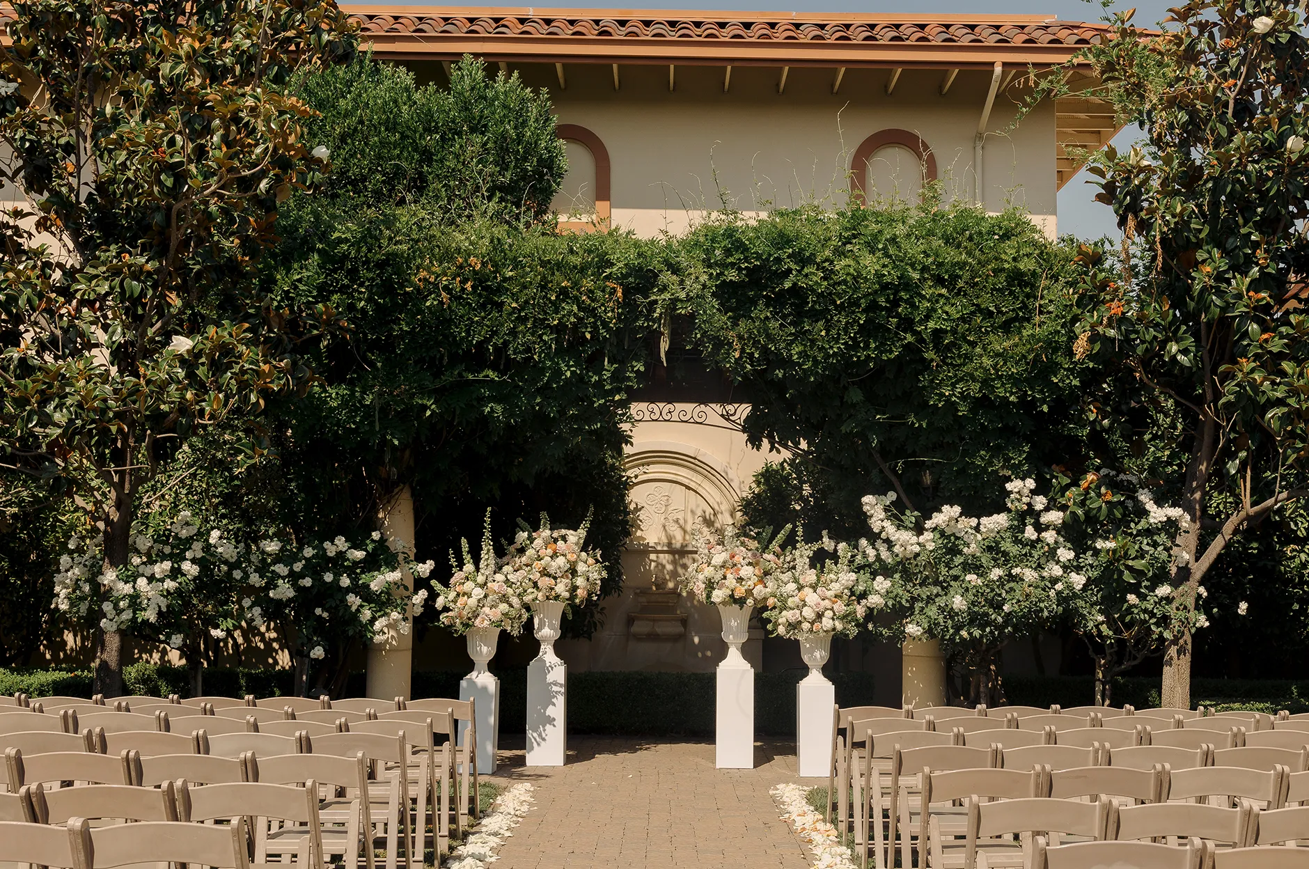 Outdoor ceremony setup at Casa Real with rows of chairs lining a petal-strewn aisle flanked by white floral arrangements in urns