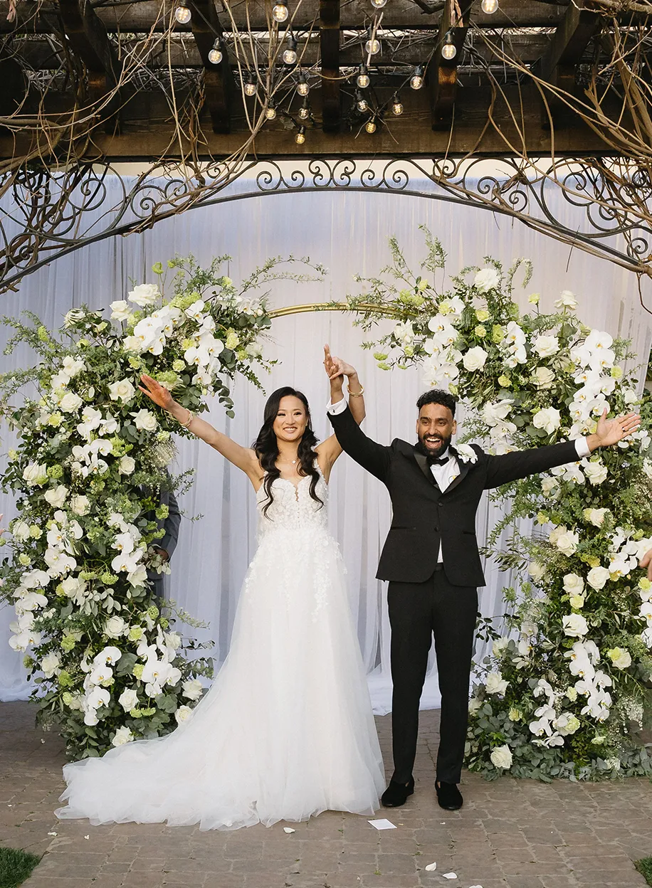 Bride and groom celebrating with arms raised beneath a lush white floral arch