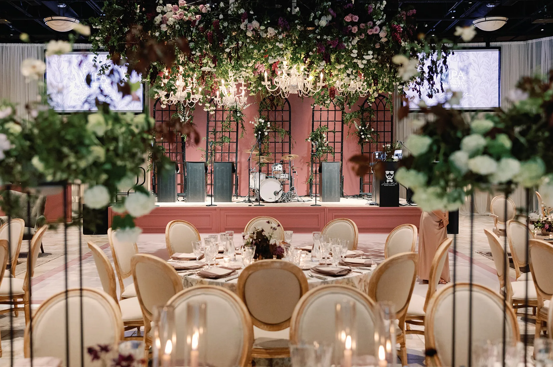Elegant banquet hall with round tables, gold chairs, a drum kit on stage, and lush floral installations hanging from the ceiling