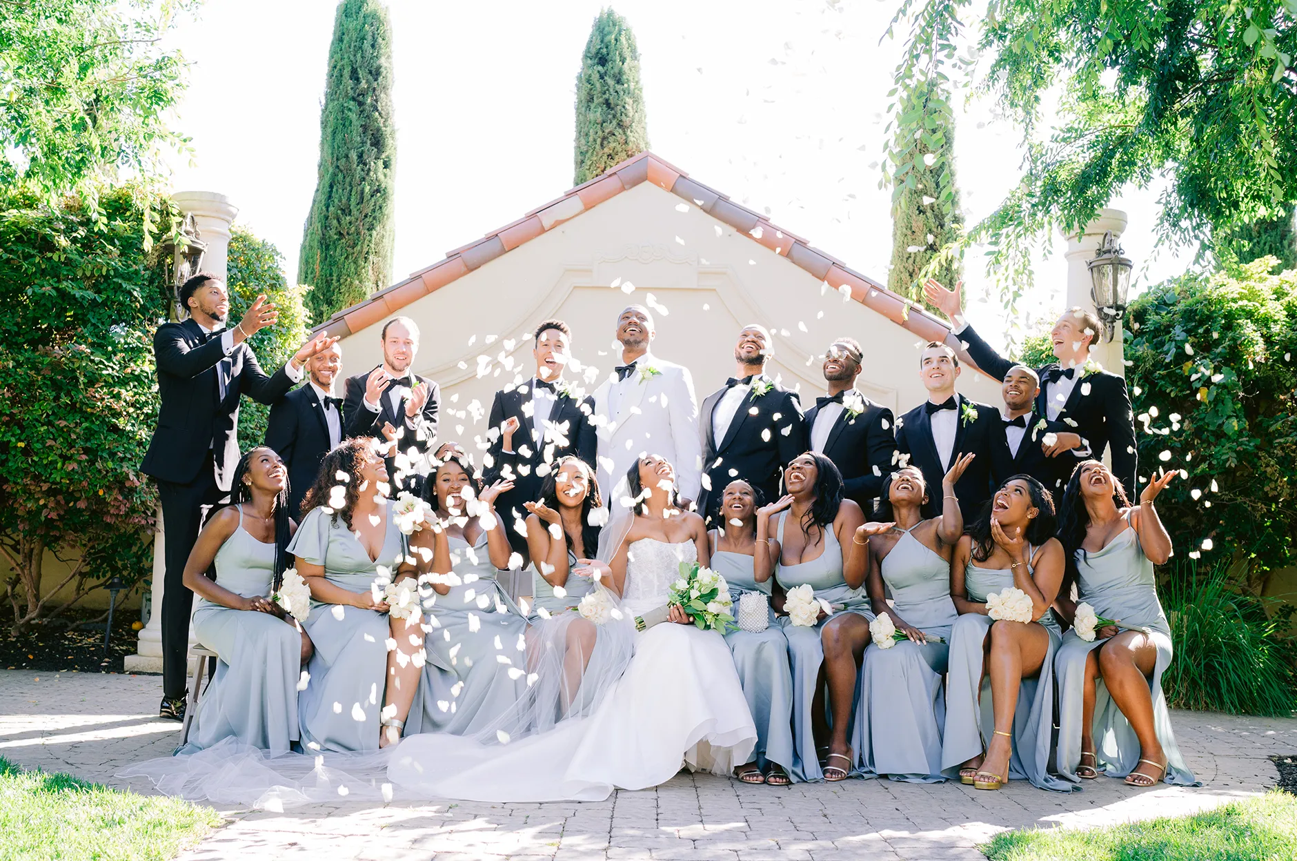 Wedding party tossing flower petals in the air in front of a vine-covered chapel with Italian cypress trees