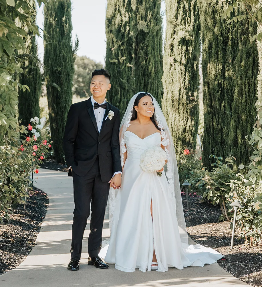 Bride and groom walking together along a garden path lined with Italian cypress trees and rose bushes