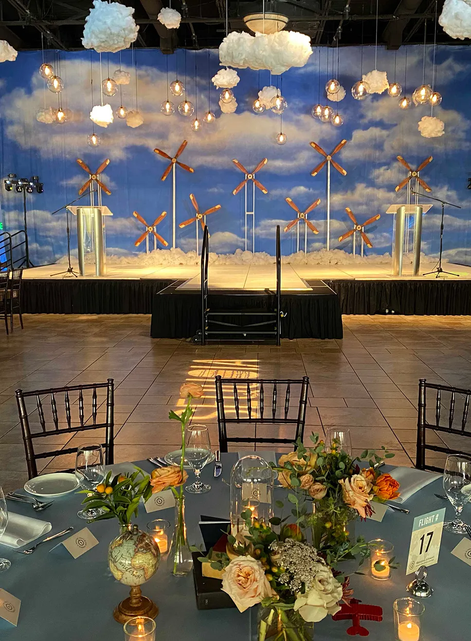 Corporate event stage with a wind turbine backdrop, cloud-covered mural, and candlelit dinner tables in the foreground