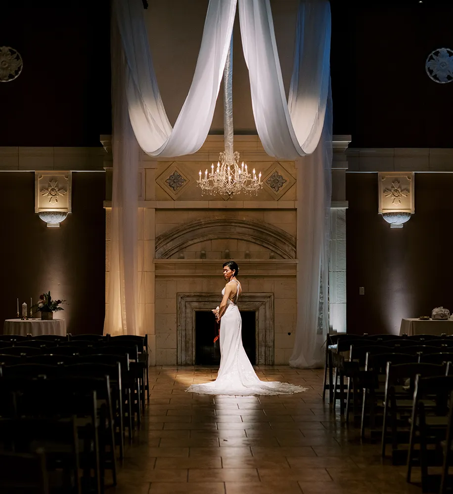 Bride standing alone in a dramatically lit ballroom with draped white fabric, a crystal chandelier, and a grand stone fireplace