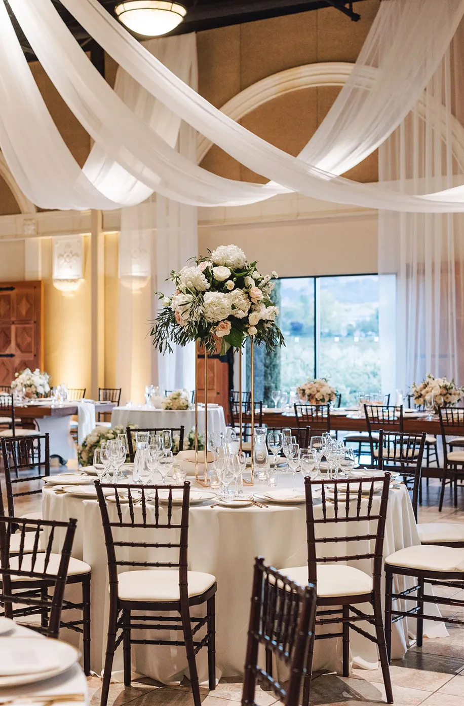 Reception tables with tall white floral centerpieces and dark chiavari chairs beneath sweeping white ceiling drapes