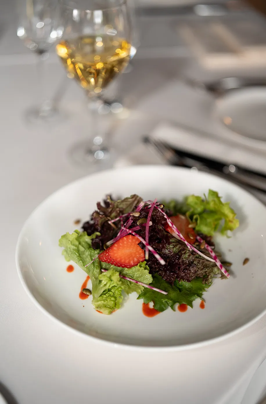 Mixed green salad with sliced strawberry and berry drizzle on a white plate beside a glass of white wine
