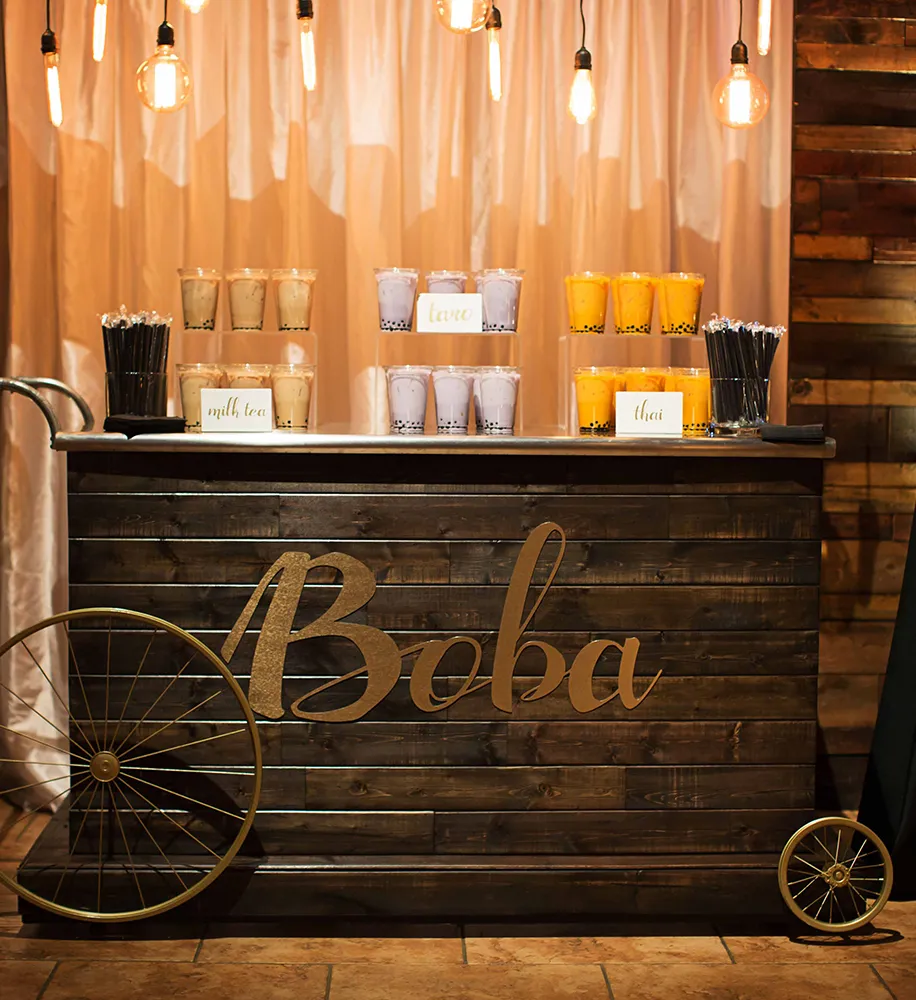 Rustic boba tea bar cart with assorted drinks displayed under warm Edison bulb lighting
