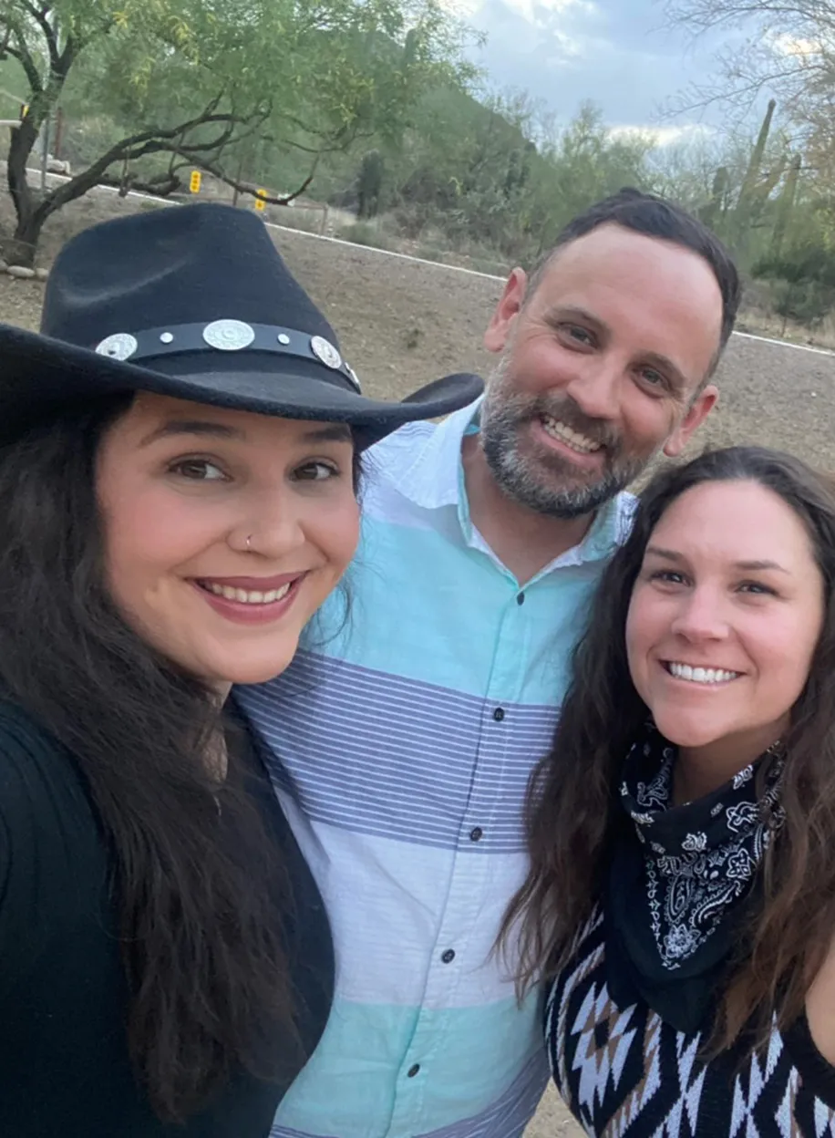Three team members in a selfie outdoors with desert greenery and a cloudy sky