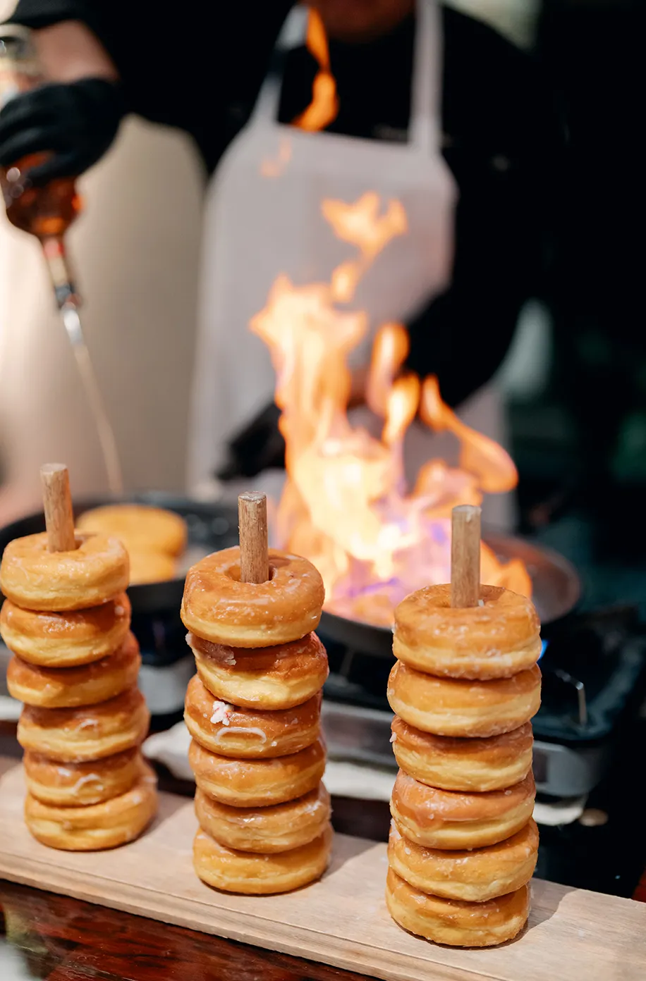Three stacks of mini doughnuts on wooden dowels being flambéed by a chef with a torch