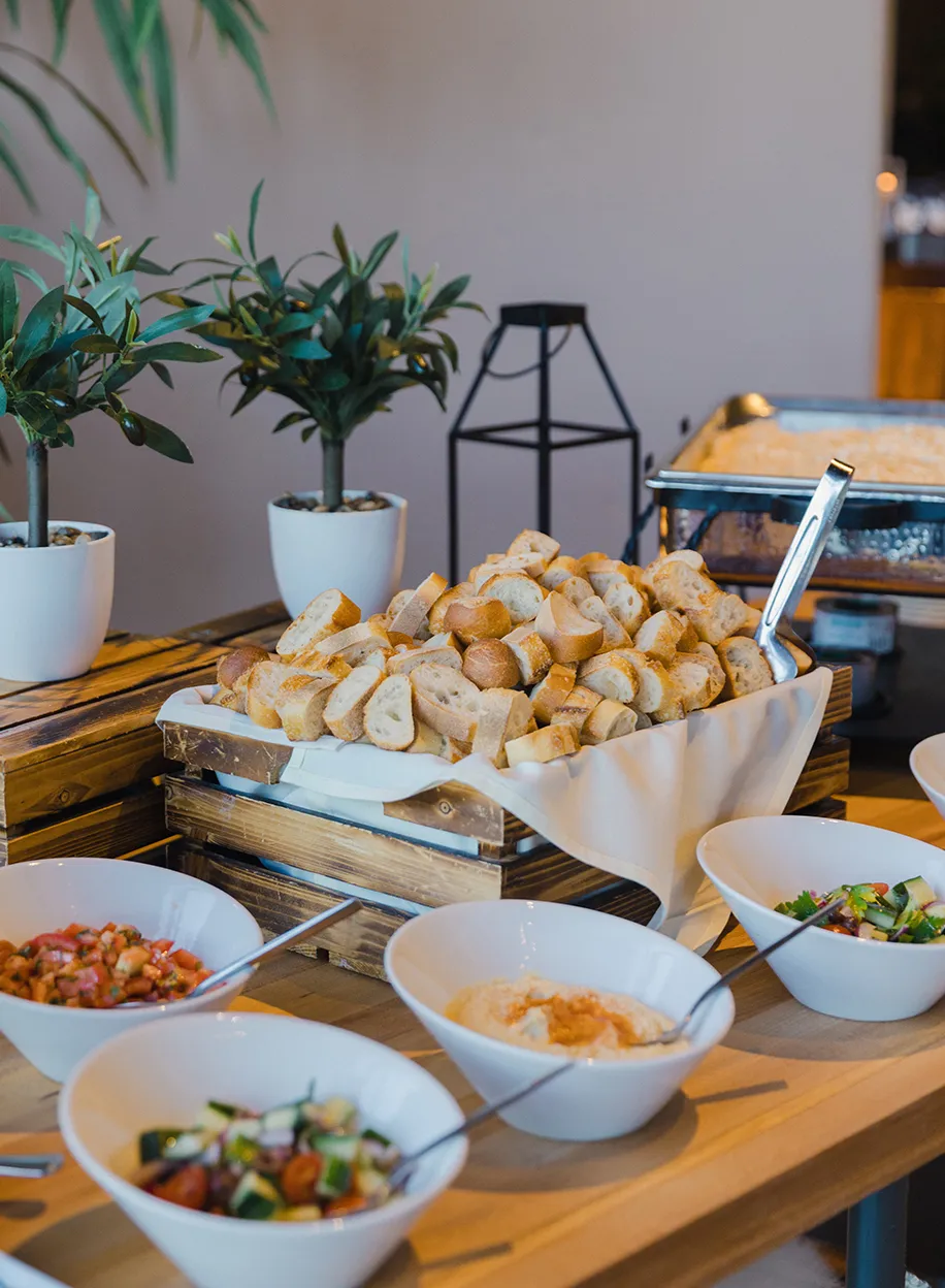 Buffet spread with baskets of bread rolls, salads, dips, and chafing dishes on a wooden table