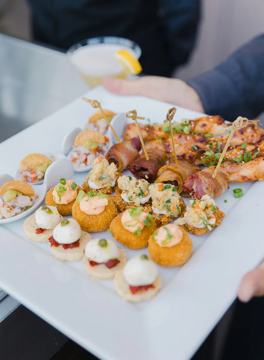 Server presenting a platter of assorted passed appetizers including bacon-wrapped bites, shrimp crostini, and sesame-glazed skewers