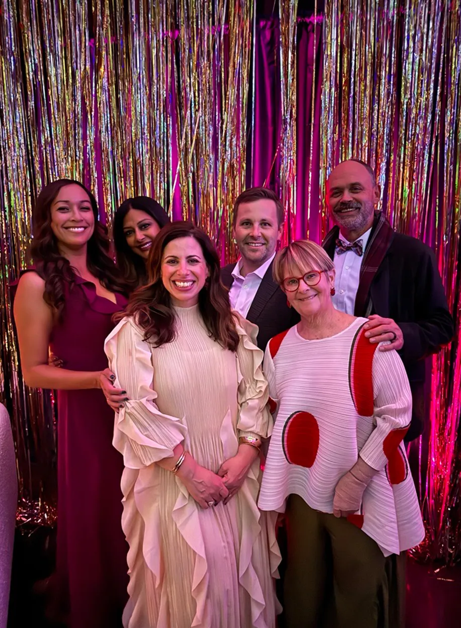 Five team members dressed up and posing together in front of a shimmering tinsel backdrop at a party