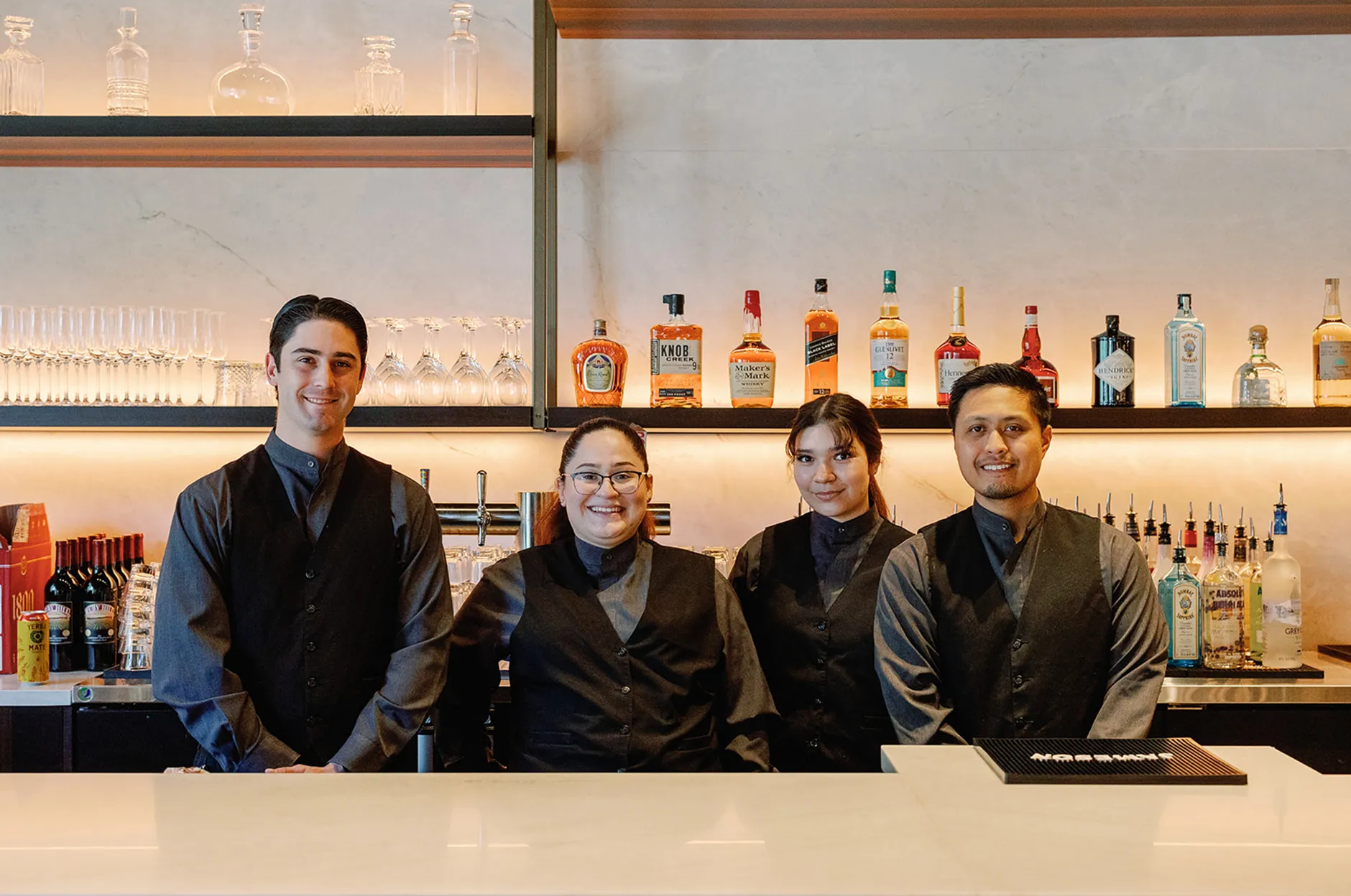 Four bartenders in black vests smiling behind the venue bar with bottles and glassware on shelves behind them