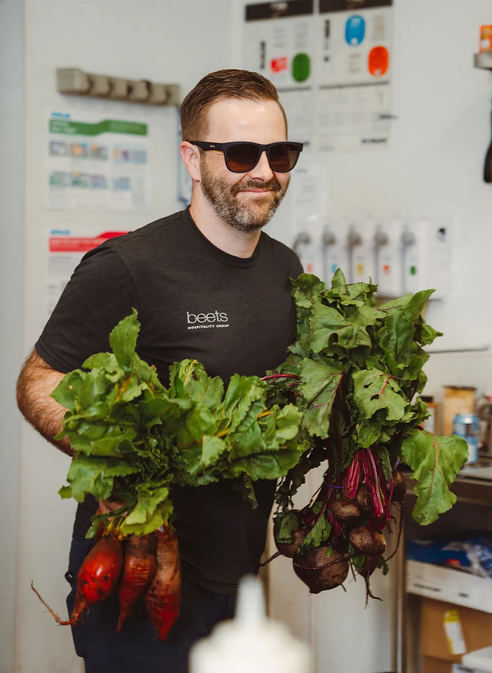 George Phillips IV in a Beets t-shirt and sunglasses holding bunches of fresh beets and greens in the kitchen