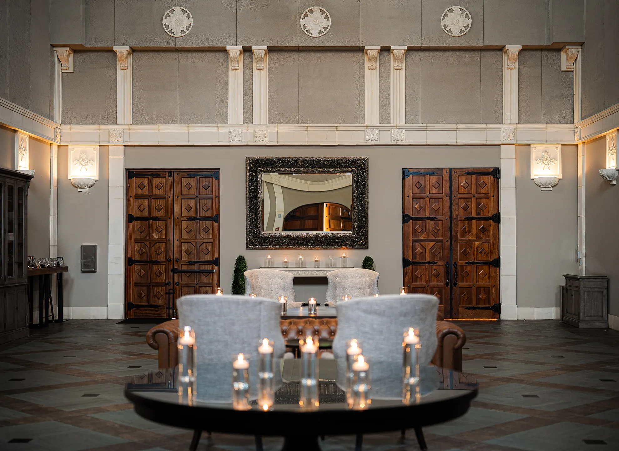 Grand entrance hall with ornate carved wooden doors, candlelit round table, upholstered chairs, and Art Deco sconces