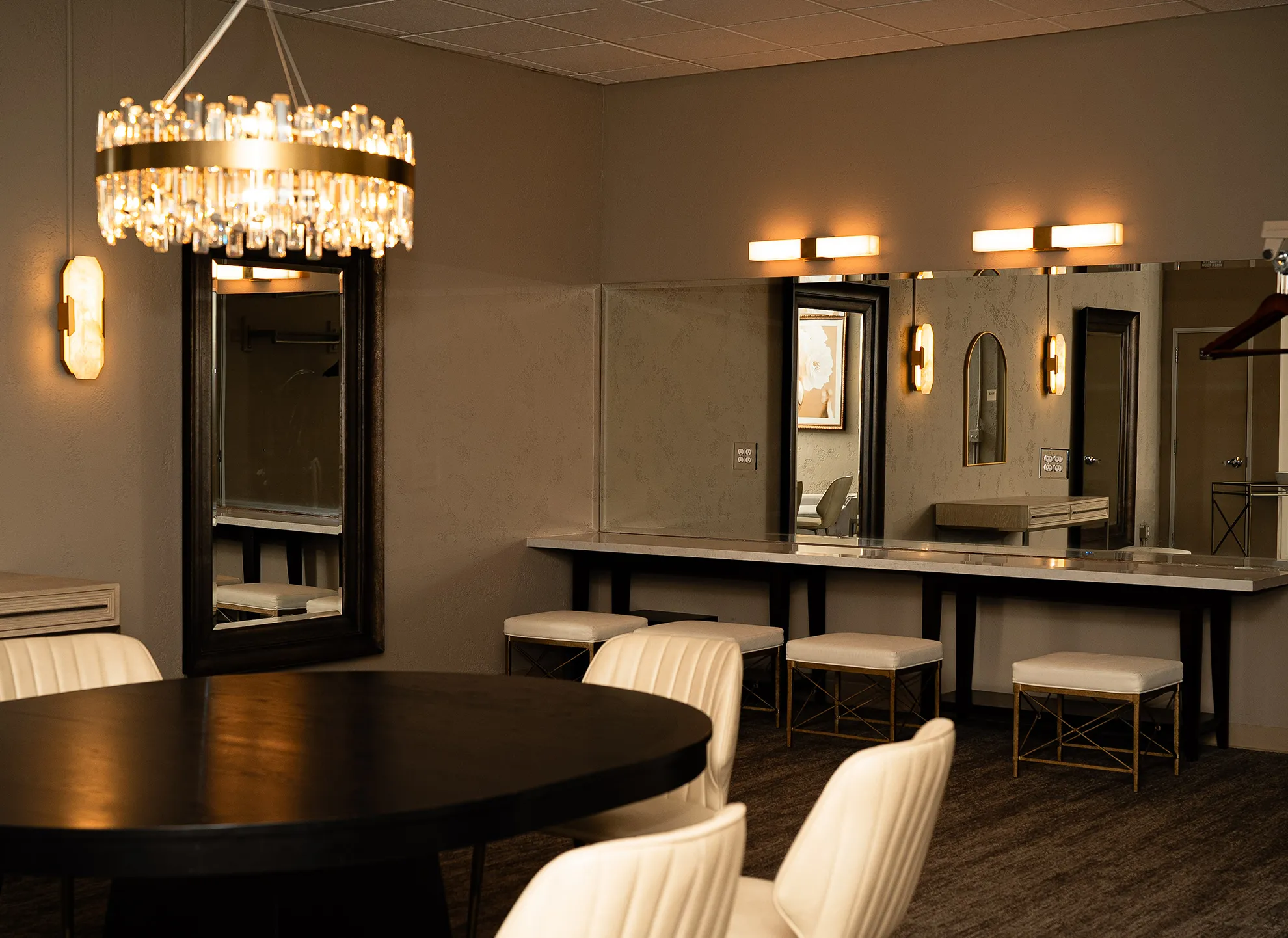 Dressing suite with a modern crystal chandelier, vanity mirrors with warm lighting, and white upholstered seating