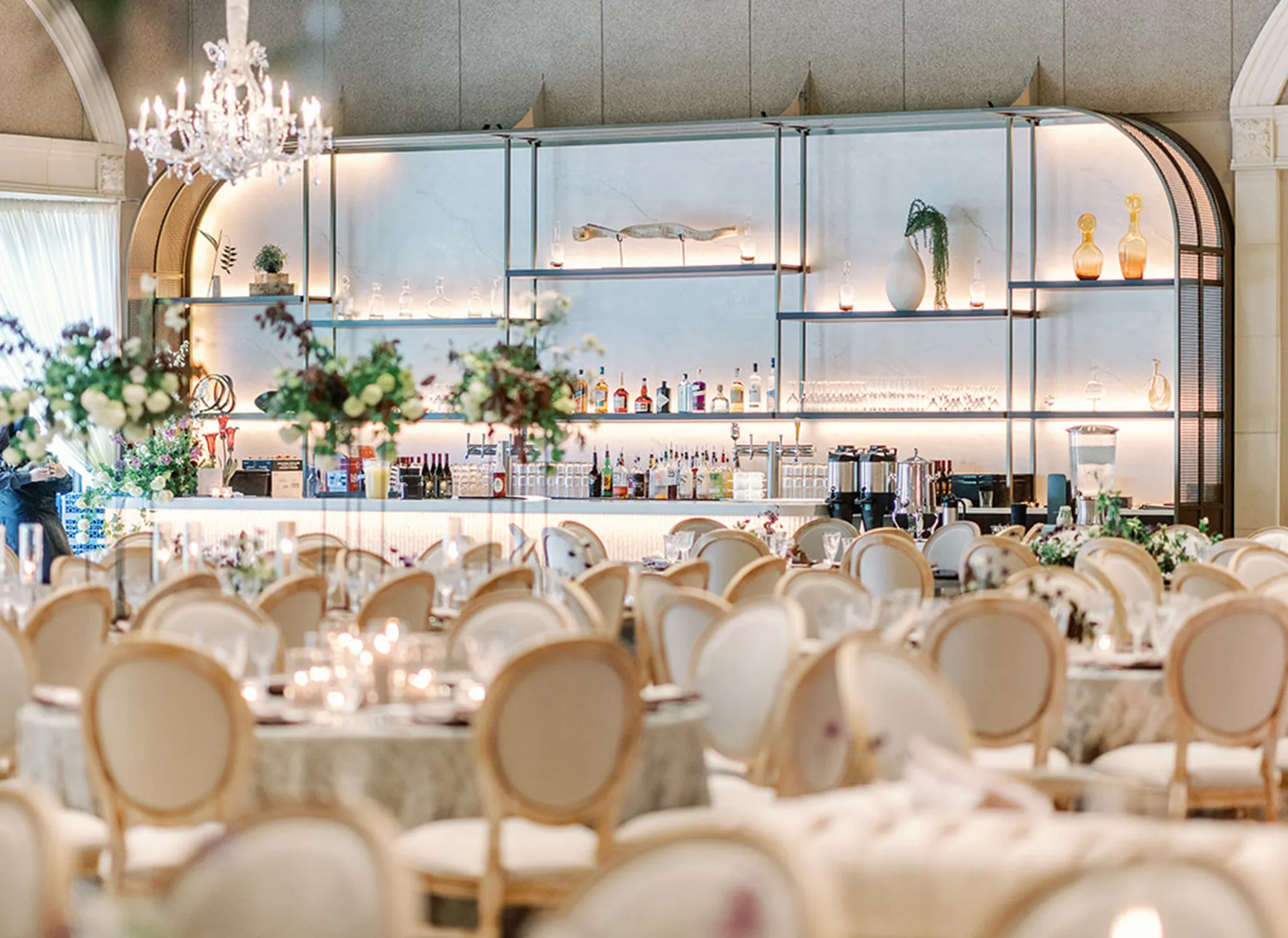 Grand Salon bar with glass shelving, crystal chandelier, and cream upholstered chairs at reception tables
