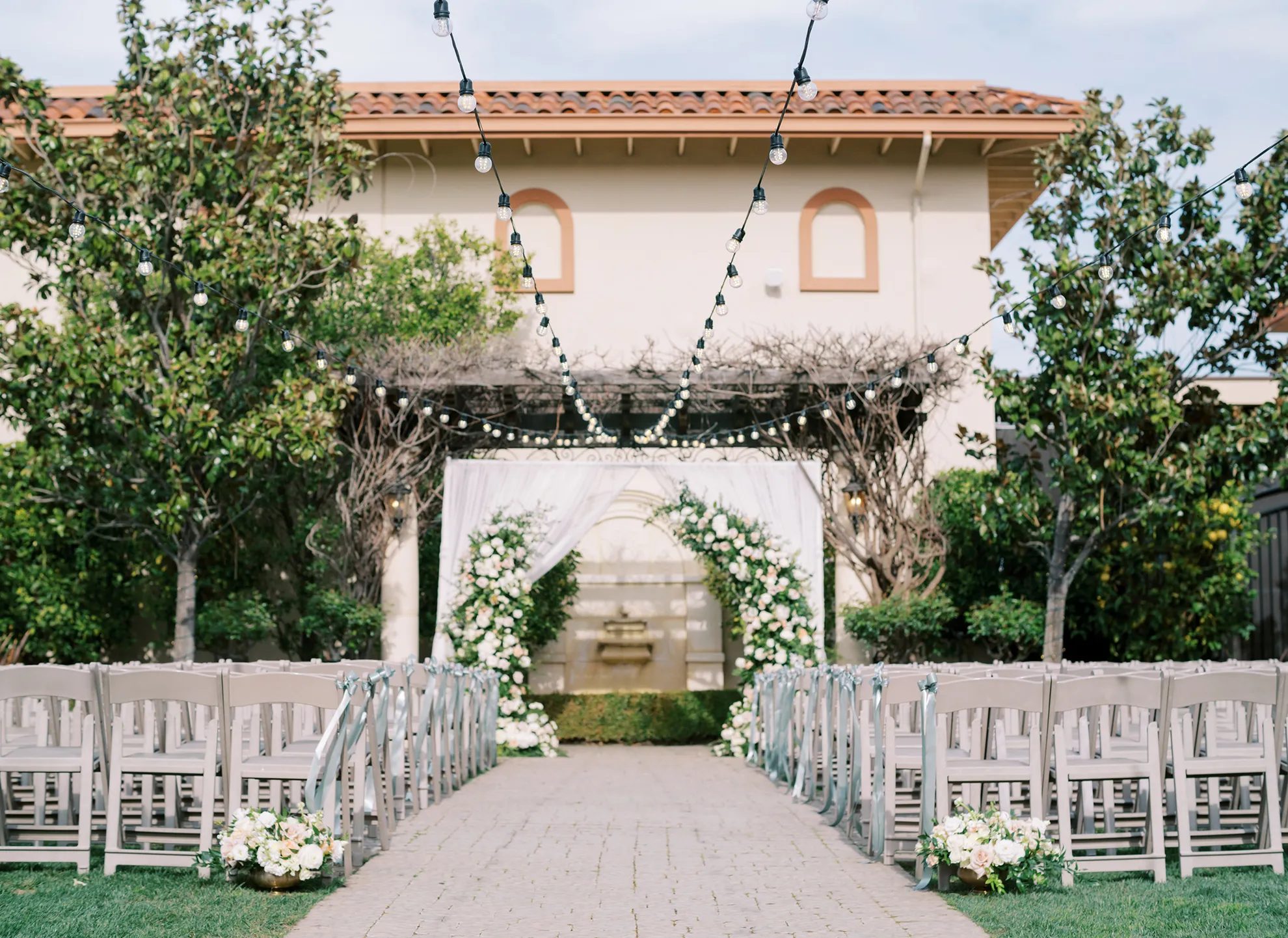 Sun Garden ceremony setup with white folding chairs, a floral-lined brick aisle, string lights, and pergola