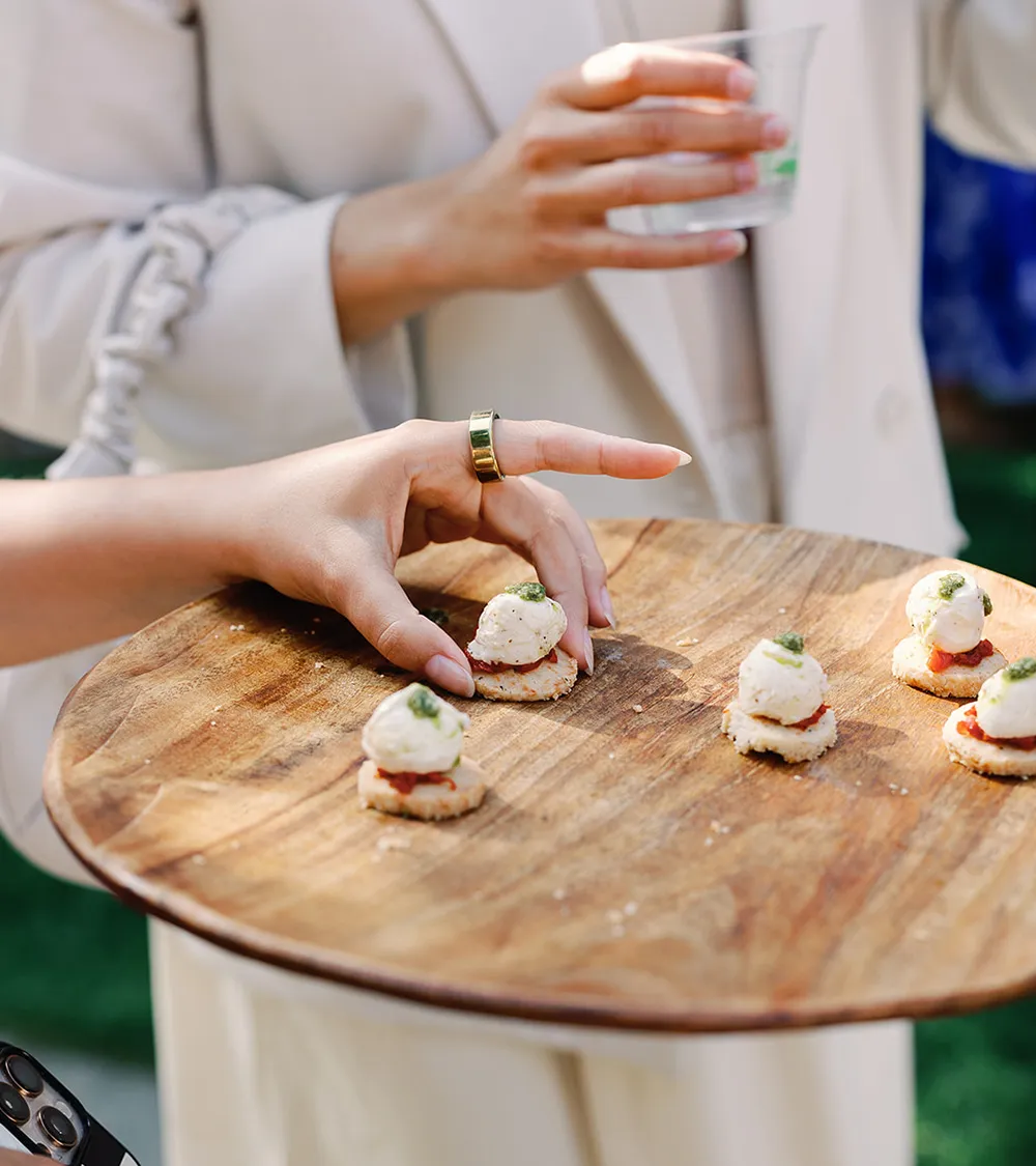 Guest selecting a canapé from a wooden serving board at an outdoor cocktail reception