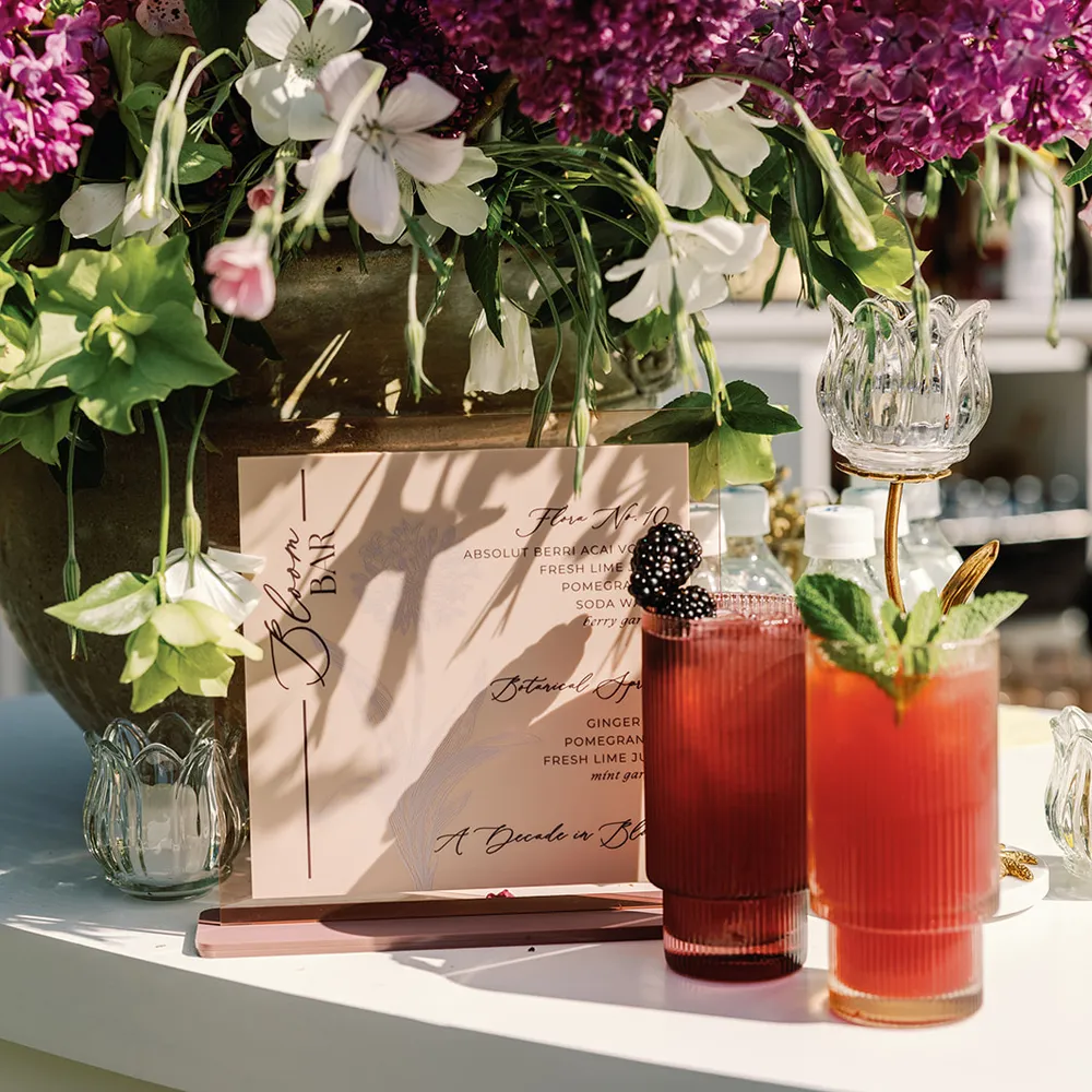 Signature cocktails in tall glasses beside a printed menu on a sunny bar top with fresh floral arrangements