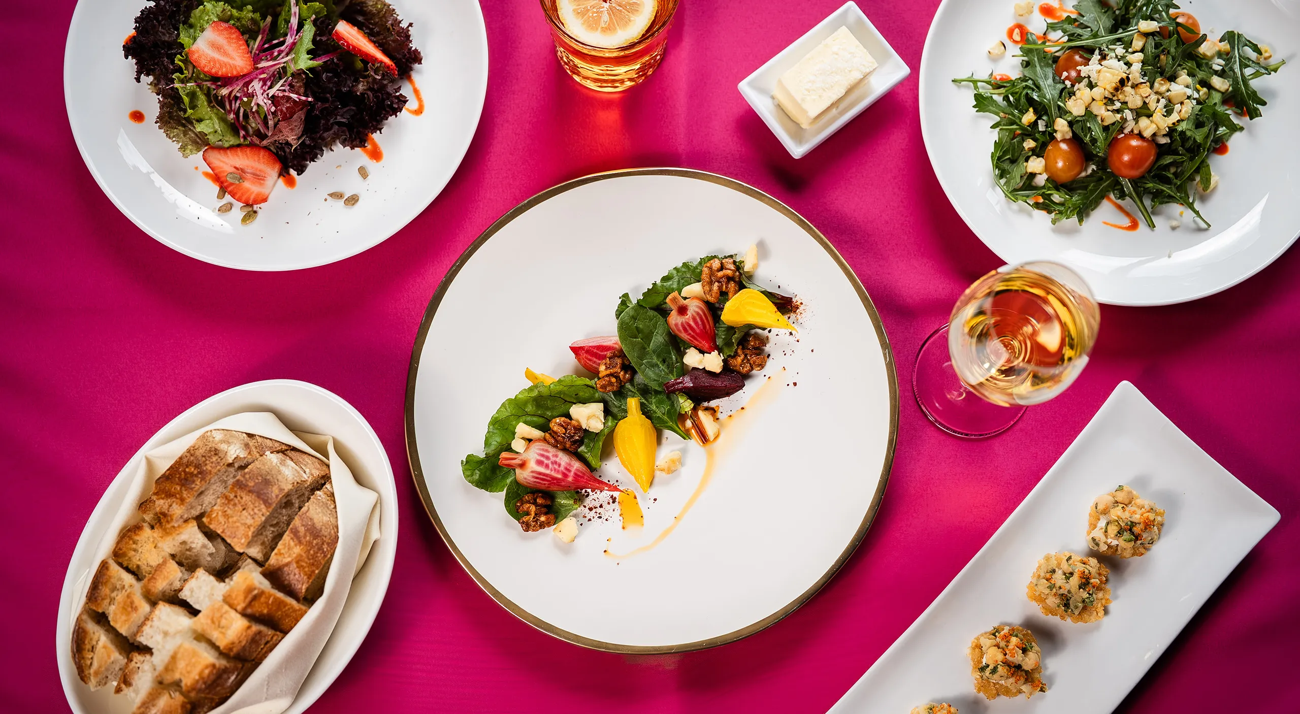 Overhead view of plated salads, artisan bread, appetizers, and rosé on a magenta linen