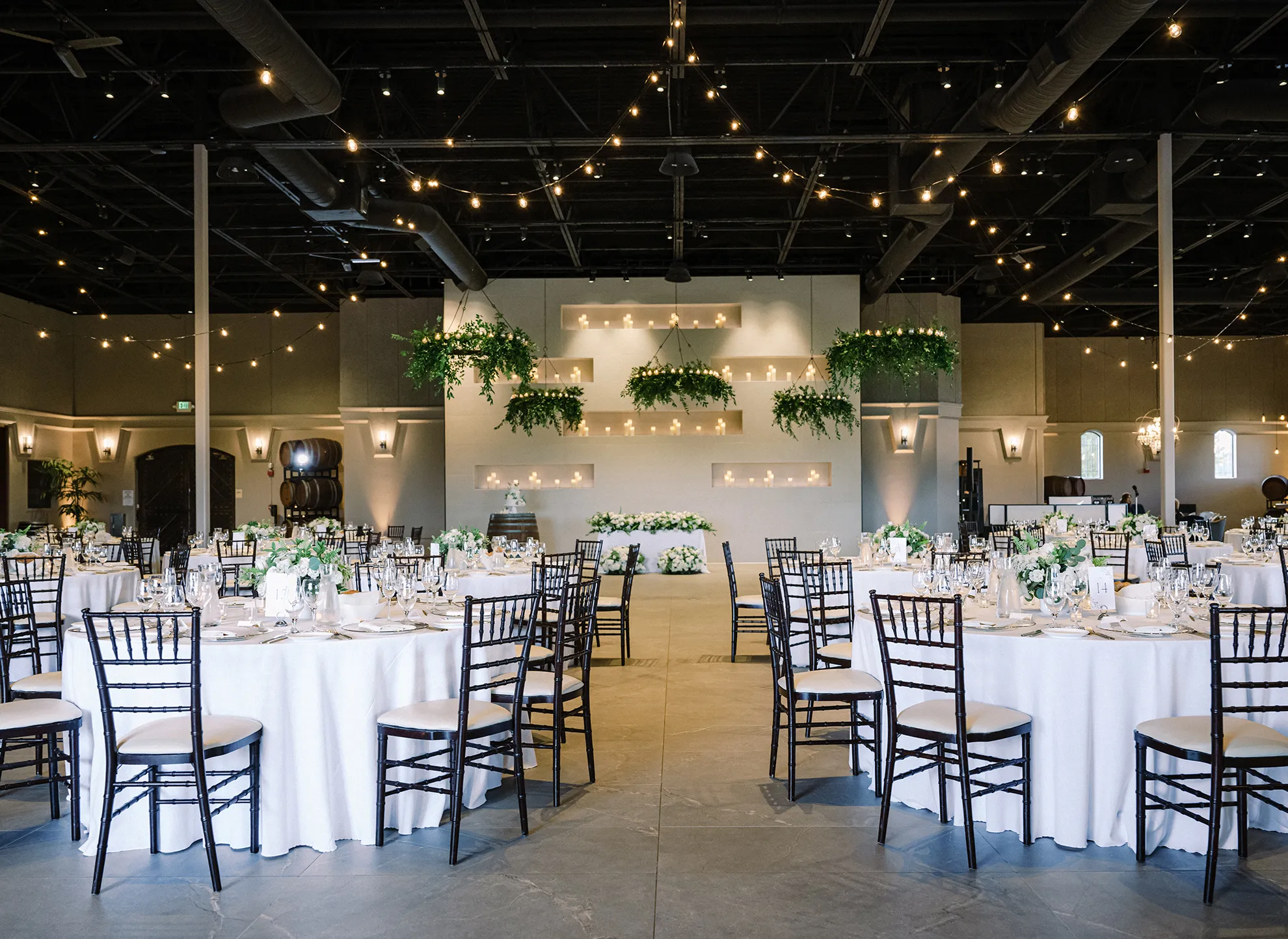 Ballroom set for a reception with round tables, black chiavari chairs, string lights, and greenery wall display