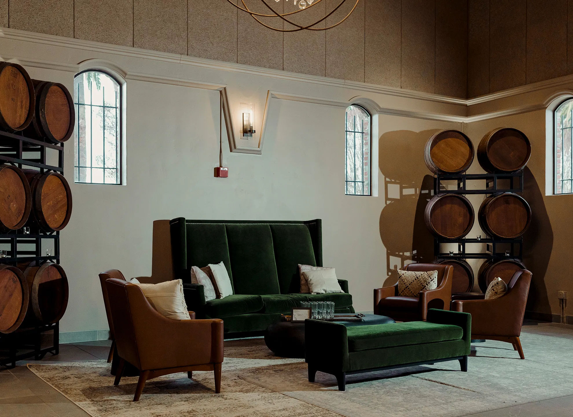 Lounge area with a green velvet sofa, leather armchairs, stacked wine barrels, and arched windows